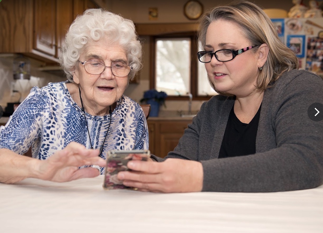 A caregiver helping an older woman with her phone at the kitchen table