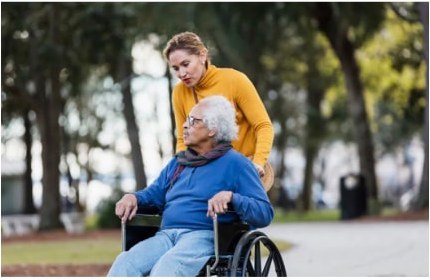 A caregiver pushing an older woman in a wheelchair through a park
