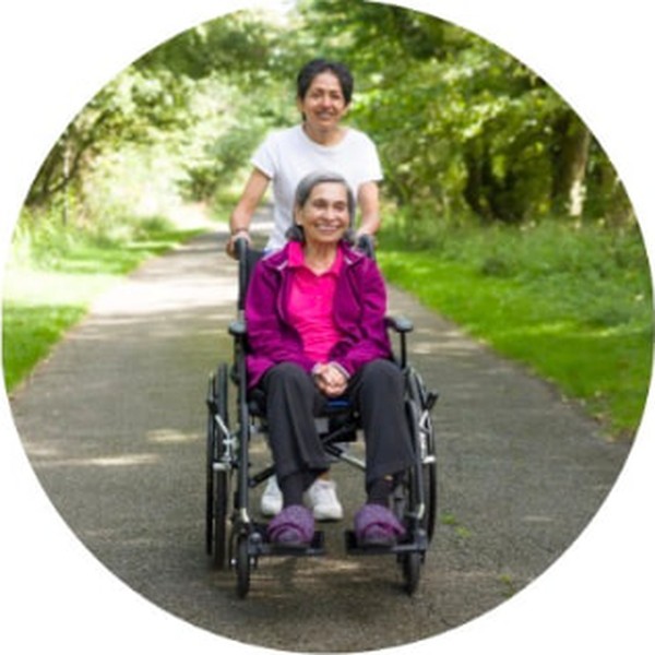 A caregiver pushing an older woman in a wheelchair through a park